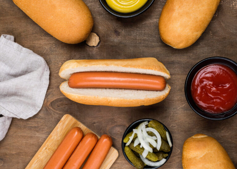 A classic Hotdog in a Hotdog Bun Recipe surrounded by condiments, pickles, onions, and additional hot dog ingredients on a wooden table.