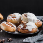 Golden brown cinnamon rolls piled on a black plate, generously topped with thick white glaze, surrounded by a cozy gray napkin and star anise