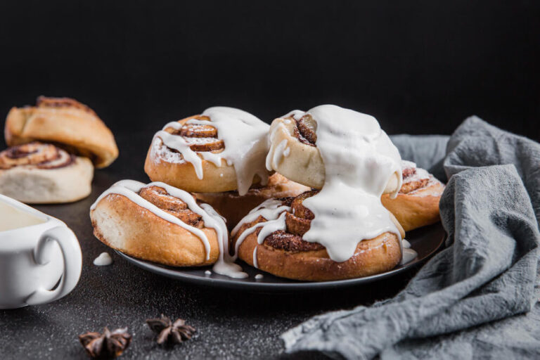 Golden brown cinnamon rolls piled on a black plate, generously topped with thick white glaze, surrounded by a cozy gray napkin and star anise