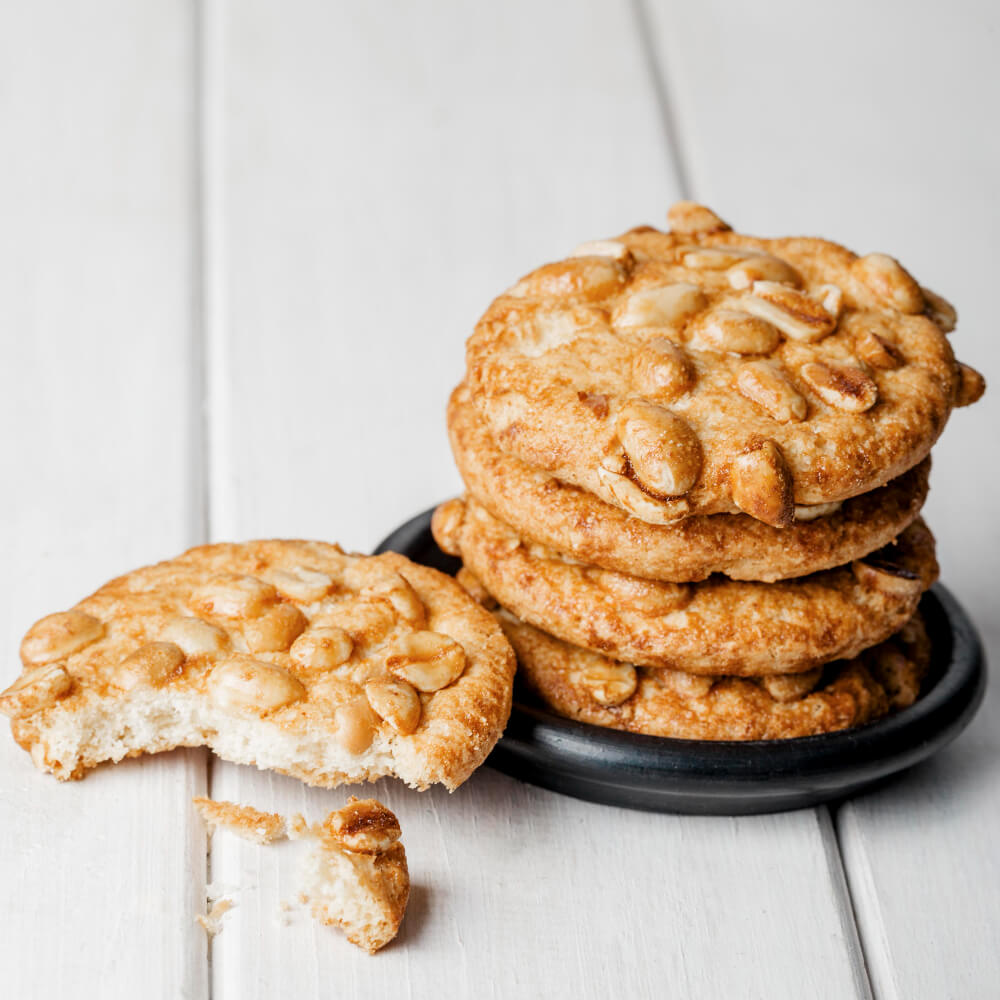 A stack of peanut cookies on a black plate with a bitten cookie and crumbs placed on a white wooden background.