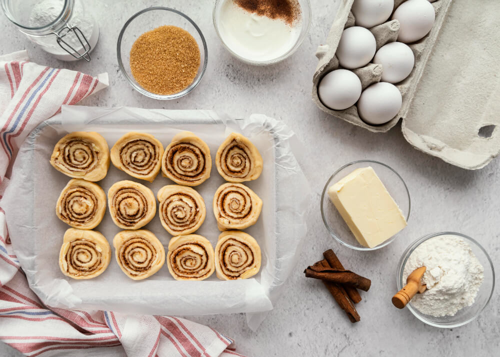 Unbaked cinnamon rolls arranged in a baking dish surrounded by baking ingredients including eggs, butter, cinnamon sticks, sugar, and flour on a light countertop.