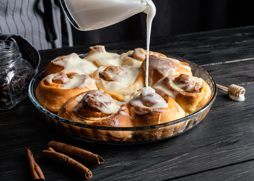 Freshly baked cinnamon rolls in a glass dish being drizzled with creamy glaze, surrounded by cinnamon sticks and honey on a dark wooden surface.