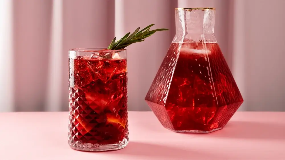 A refreshing Aperol Spritz served in a diamond-patterned glass with a rosemary garnish, paired with a matching faceted carafe on a pink surface.