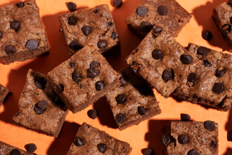 Freshly baked chocolate chip brownie squares scattered on an orange surface with chocolate chips sprinkled around.