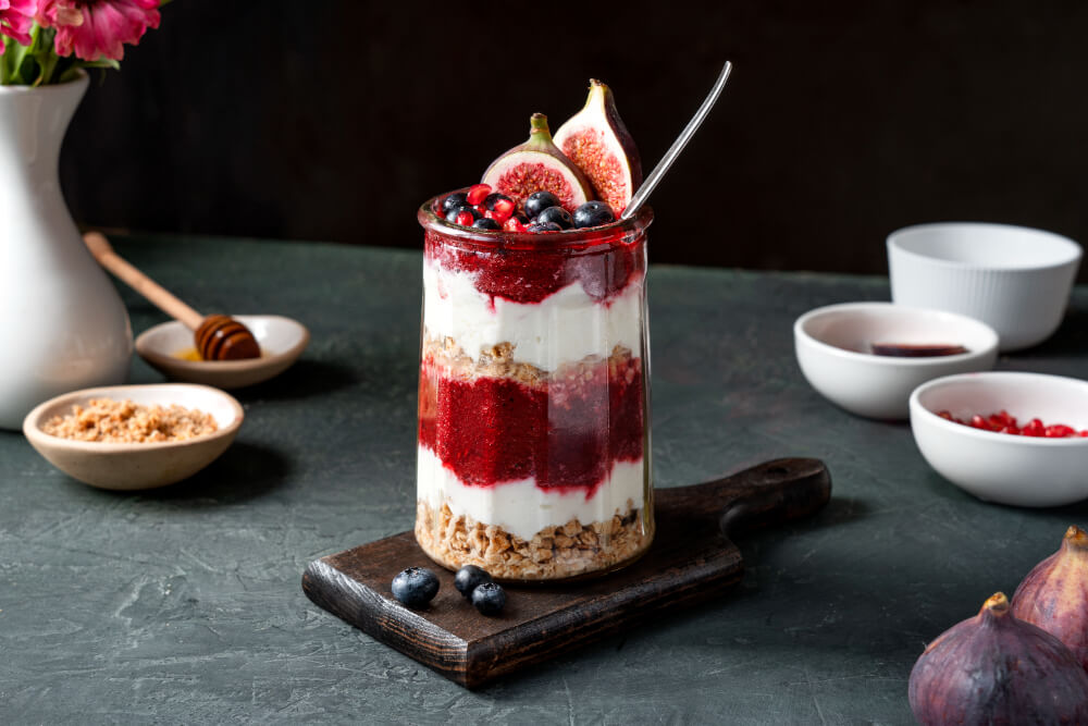 A gourmet yogurt parfait served in a tall glass, layered with granola, fresh figs, berry compote, and topped with blueberries and a fig slice, presented on a wooden board