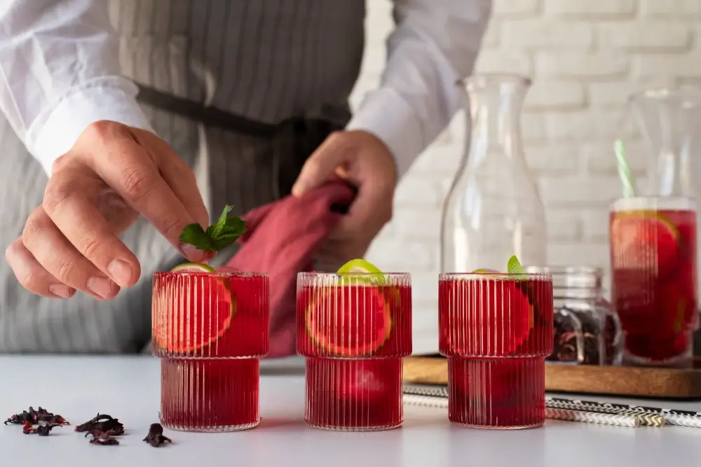 A bartender garnishing a rAperol Spritz with fresh mint leaves in stylish glasses, accompanied by lime slices and hibiscus petals on a white countertop.