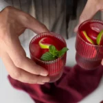 Close-up of hands holding two ribbed glasses filled with a vibrant Aperol Spritz, garnished with mint leaves and lime slices on a white countertop.