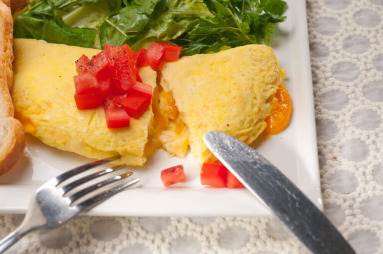 A cheese omelette topped with fresh diced tomatoes, served with arugula salad and toasted bread on a white plate, partially cut to reveal the melted cheese inside.