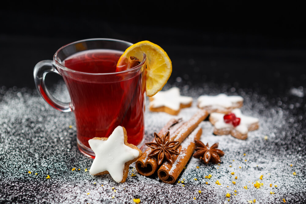 A glass mug of non-alcoholic Christmas punch garnished with a lemon slice and cinnamon stick, surrounded by star-shaped cookies, cinnamon, and star anise on a snowy surface.