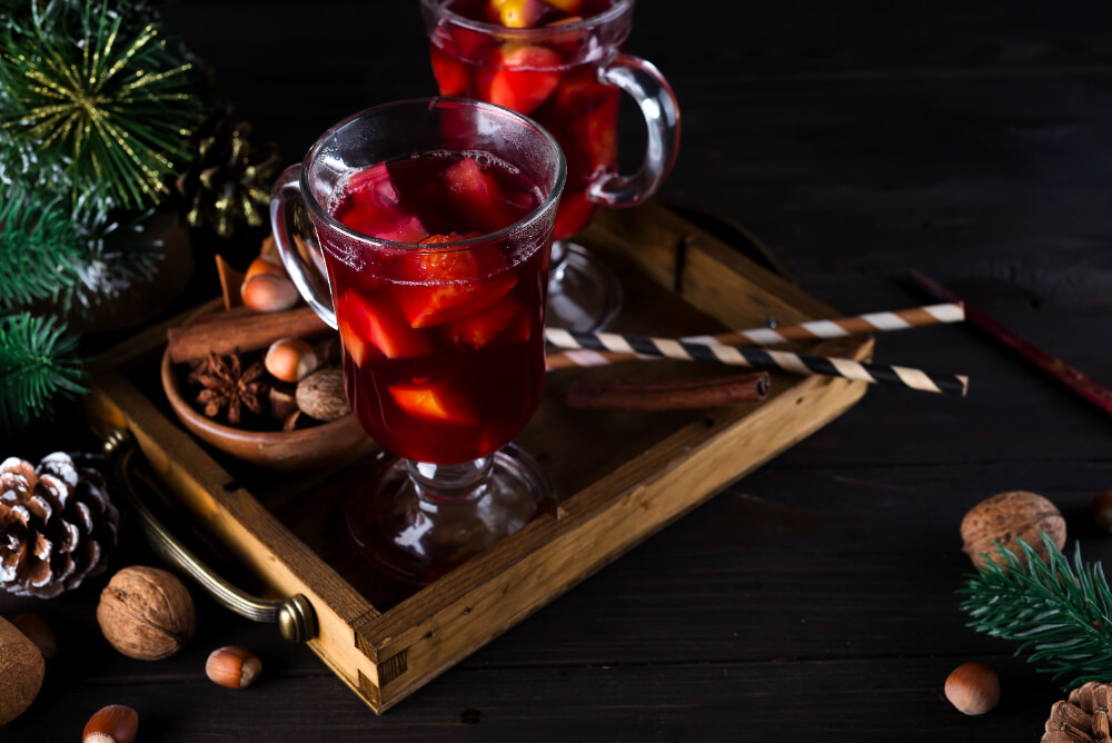 A glass mug of festive non-alcoholic Christmas punch with fruit pieces, cinnamon sticks, and holiday decorations on a wooden tray.