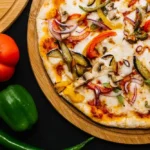 "Homemade veggie pizza on a wooden board with fresh toppings of bell peppers, red onions, mushrooms, and tomatoes alongside fresh vegetables."