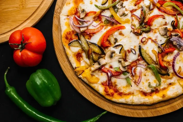 "Homemade veggie pizza on a wooden board with fresh toppings of bell peppers, red onions, mushrooms, and tomatoes alongside fresh vegetables."