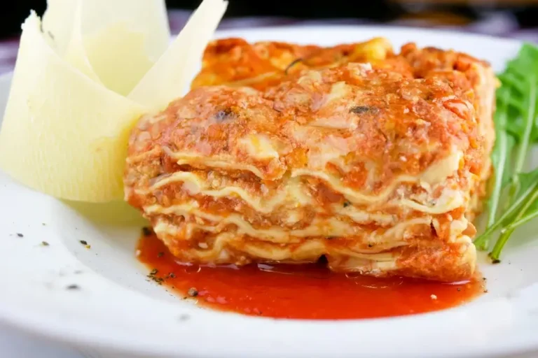 A slice of lasagna on a white plate, topped with rich tomato sauce and garnished with fresh greens and shaved cheese.