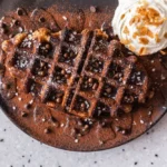 "Delicious chocolate waffle topped with chocolate chips, whipped cream, cocoa powder, and a sprinkle of sugar served on a black plate."