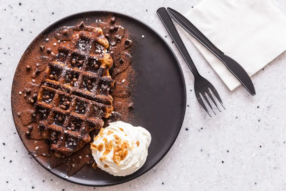 "Chocolate waffle served on a black plate with chocolate chips, cocoa powder, whipped cream, and a sprinkle of sugar alongside a fork and knife."