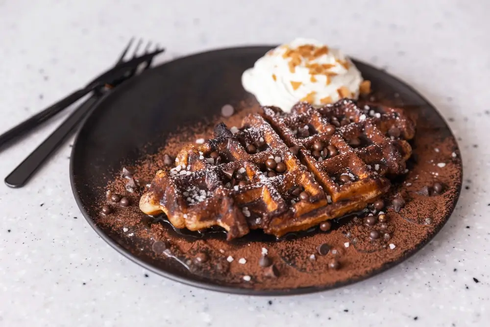 "Close-up of a chocolate waffle topped with chocolate chips, cocoa powder, whipped cream, and a sprinkle of sugar served on a black plate."