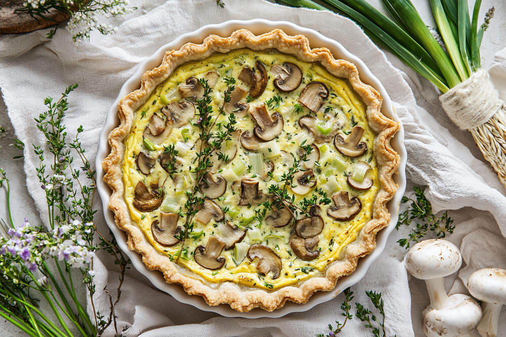 A freshly baked mushroom quiche in a golden, flaky crust, garnished with sprigs of fresh thyme and surrounded by mushrooms and green onions.