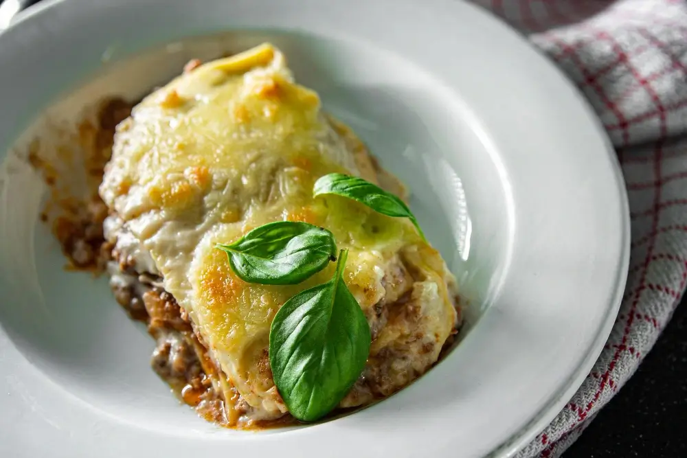 A slice of creamy lasagna served on a white plate, topped with melted cheese and fresh basil leaves.