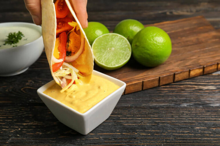 A fish taco filled with vegetables dipped into a bowl of creamy yellow sauce, with fresh limes and a wooden board in the background on a rustic dark wooden table.