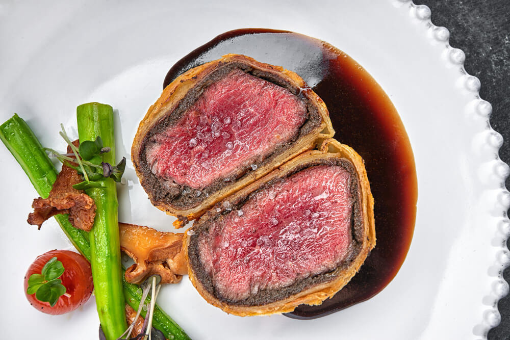 Plate of Individual Beef Wellington served with asparagus, cherry tomato, and a rich sauce.