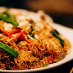 A plate of golden stir-fried noodles garnished with green onions and red chilies.