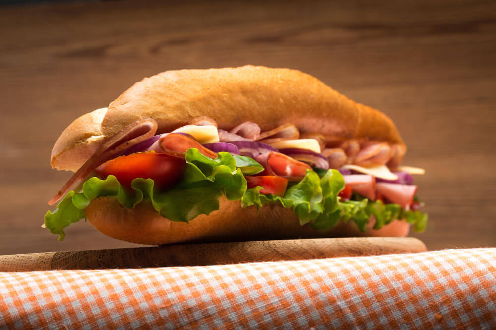 A classic grinder sandwich filled with fresh lettuce, tomatoes, deli meats, and sliced cheese, placed on a wooden surface with a gingham-patterned napkin.