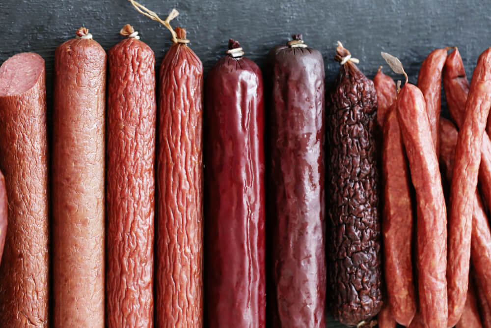 An assortment of sausages displayed in various sizes and textures, including smoked and cured varieties, arranged on a dark surface.