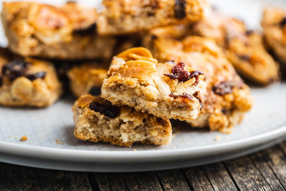 A close-up of nut and cranberry cookie bars stacked on a plate, with visible almonds, dried cranberries, and a golden, crumbly texture.