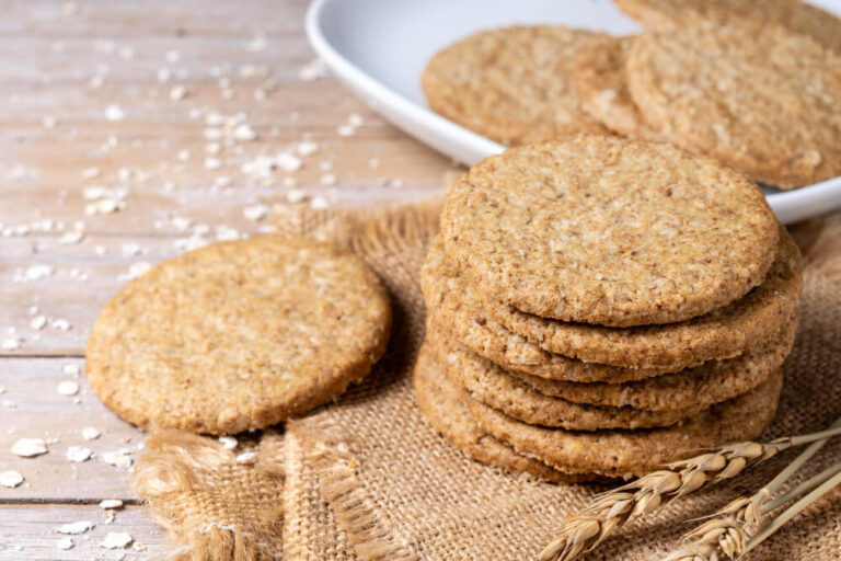 A stack of golden-brown oatmeal cookies on burlap fabric with one cookie leaning in the foreground, oats scattered on a wooden surface.