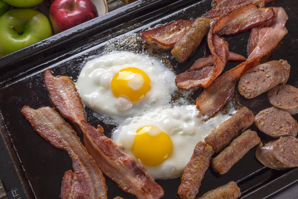 Two sunny-side-up eggs with crispy bacon, sausage links, and sliced sausage on a black griddle.