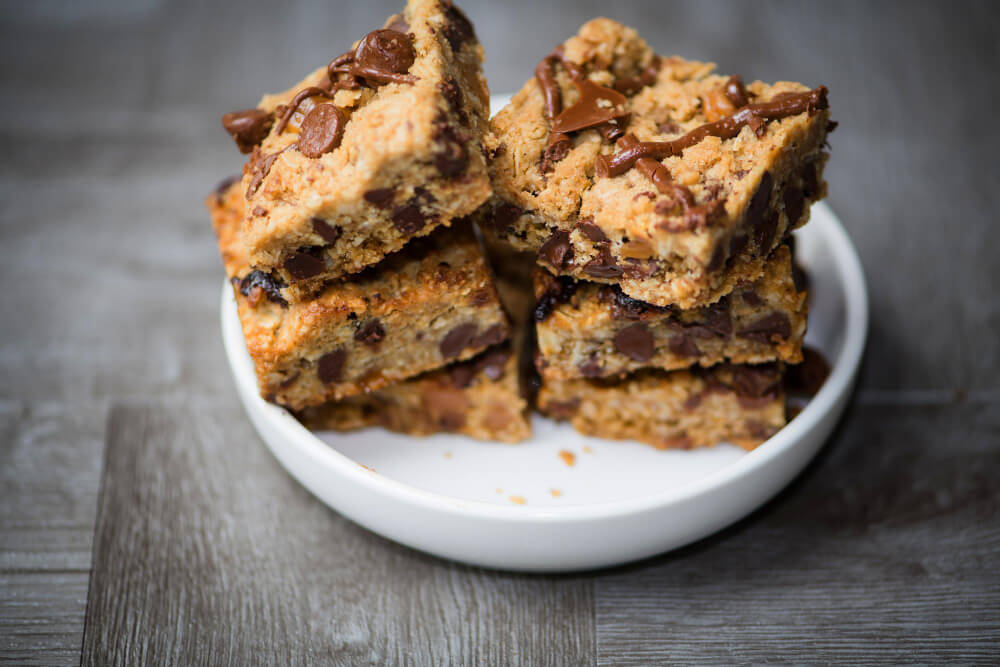 A stack of freshly baked chocolate chip cookie bars drizzled with chocolate, placed on a white plate against a rustic wooden background.