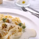 A plate of creamy chicken fettuccine Alfredo garnished with broccoli, served on a white dish with cutlery in the background.