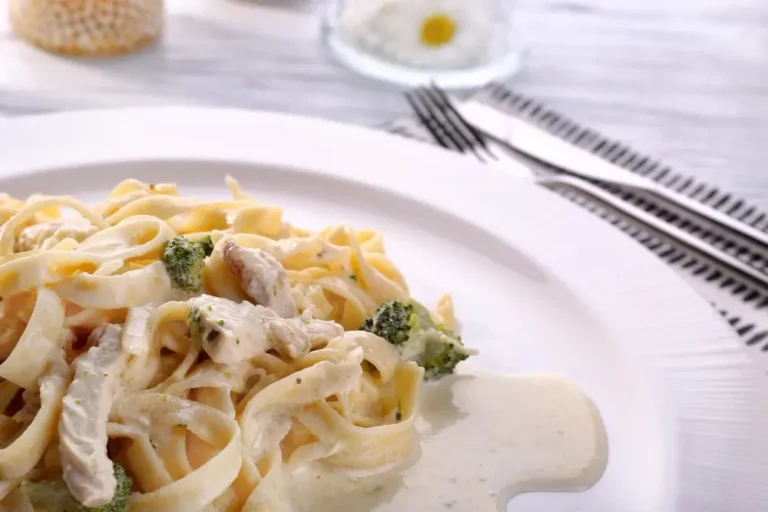 A plate of creamy chicken fettuccine Alfredo garnished with broccoli, served on a white dish with cutlery in the background.