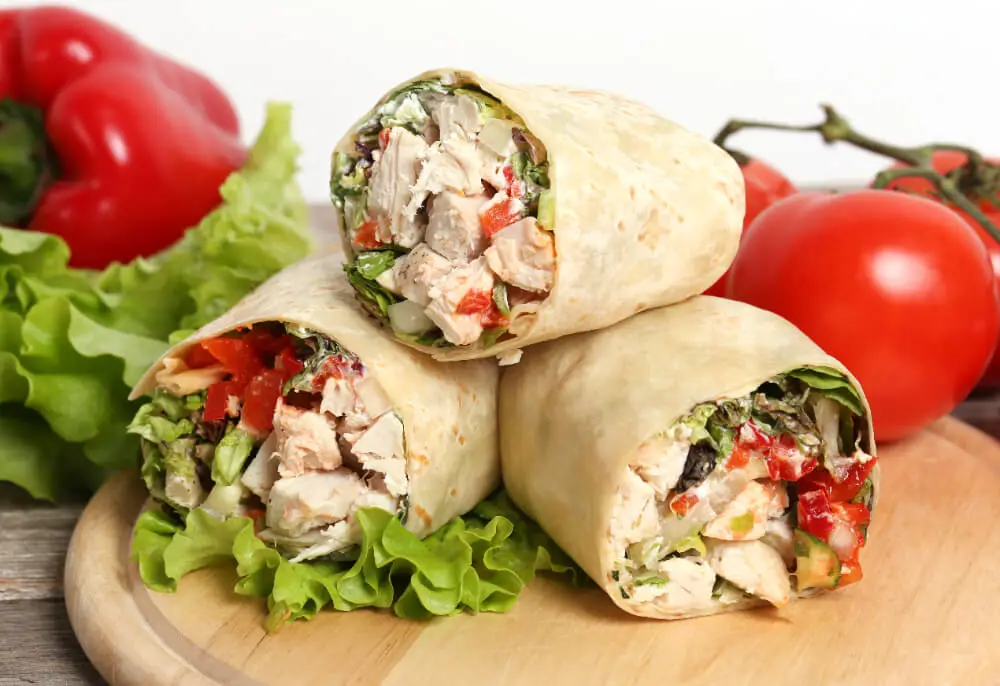 Stacked Chicken Caesar Wraps filled with chicken, lettuce, and colorful vegetables, surrounded by fresh tomatoes and bell peppers.