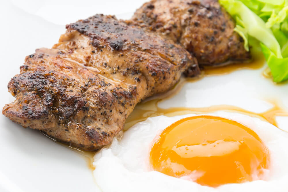 Grilled steak with a sunny-side-up egg served on a white plate, garnished with fresh green lettuce.