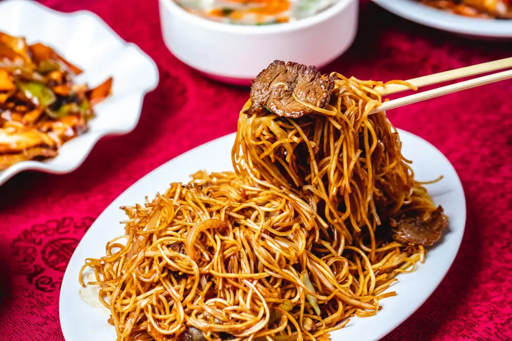 A plate of stir-fried noodles with slices of beef, served with chopsticks and accompanied by side dishes on a red tablecloth.