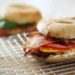 "Bagel sandwich with bacon, egg, cheese, and tomato on a cooling rack."