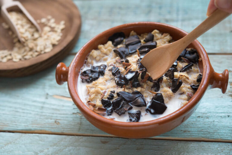 A rustic bowl of creamy oatmeal topped with dark chocolate chunks and shredded coconut, with a wooden spoon stirring, set on a light blue wooden surface.