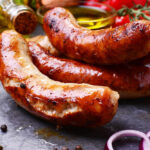 Grilled sausages served on a slate platter, surrounded by fresh ingredients like tomatoes, onions, spices, and olive oil.