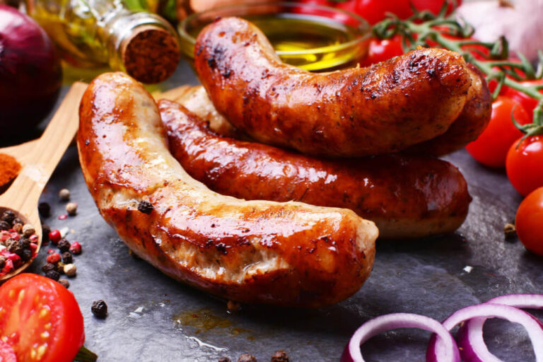 Grilled sausages served on a slate platter, surrounded by fresh ingredients like tomatoes, onions, spices, and olive oil.