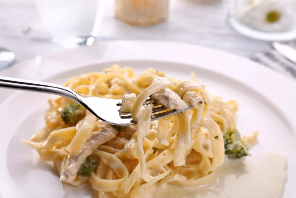 A fork lifting a bite of creamy chicken fettuccine Alfredo garnished with broccoli, served on a white plate.