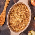 Freshly baked apple crumble in a ceramic dish surrounded by sliced apples and wooden utensils on a rustic countertop.