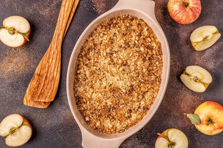 Freshly baked apple crumble in a ceramic dish surrounded by sliced apples and wooden utensils on a rustic countertop.