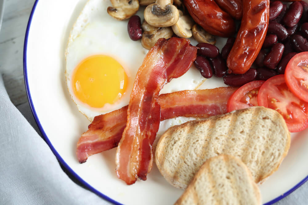 A traditional English breakfast with fried egg, crispy bacon, grilled sausages, sautéed mushrooms, kidney beans, sliced tomatoes, and toasted bread.