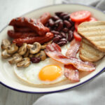 A traditional English breakfast plate with fried egg, crispy bacon, grilled sausages, sautéed mushrooms, kidney beans, sliced tomatoes, and toasted bread.