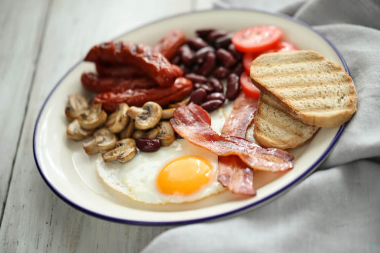 A traditional English breakfast plate with fried egg, crispy bacon, grilled sausages, sautéed mushrooms, kidney beans, sliced tomatoes, and toasted bread.