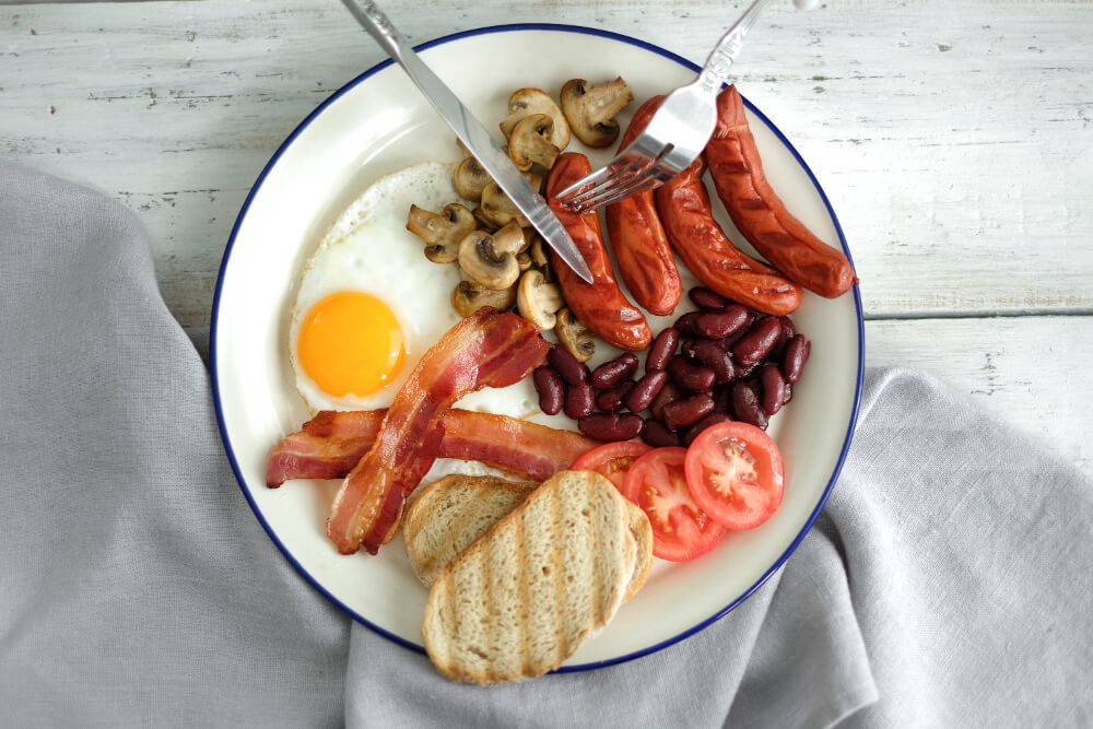 A traditional English breakfast with fried egg, crispy bacon, grilled sausages, sautéed mushrooms, kidney beans, sliced tomatoes, toasted bread, and utensils.