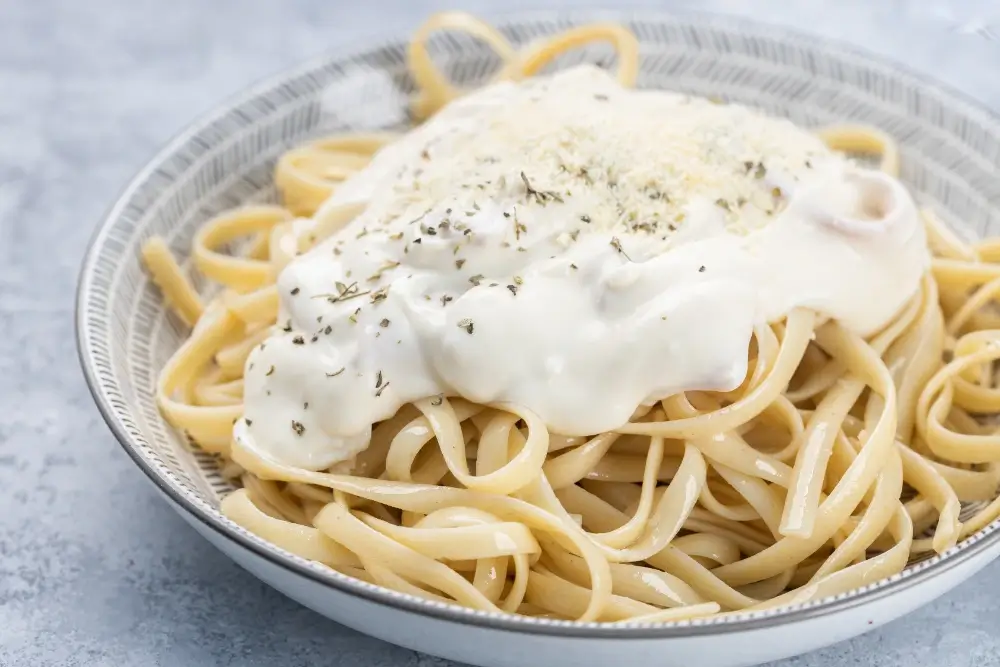 A bowl of fettuccine pasta topped with creamy Alfredo sauce and garnished with a sprinkle of herbs and grated Parmesan cheese.