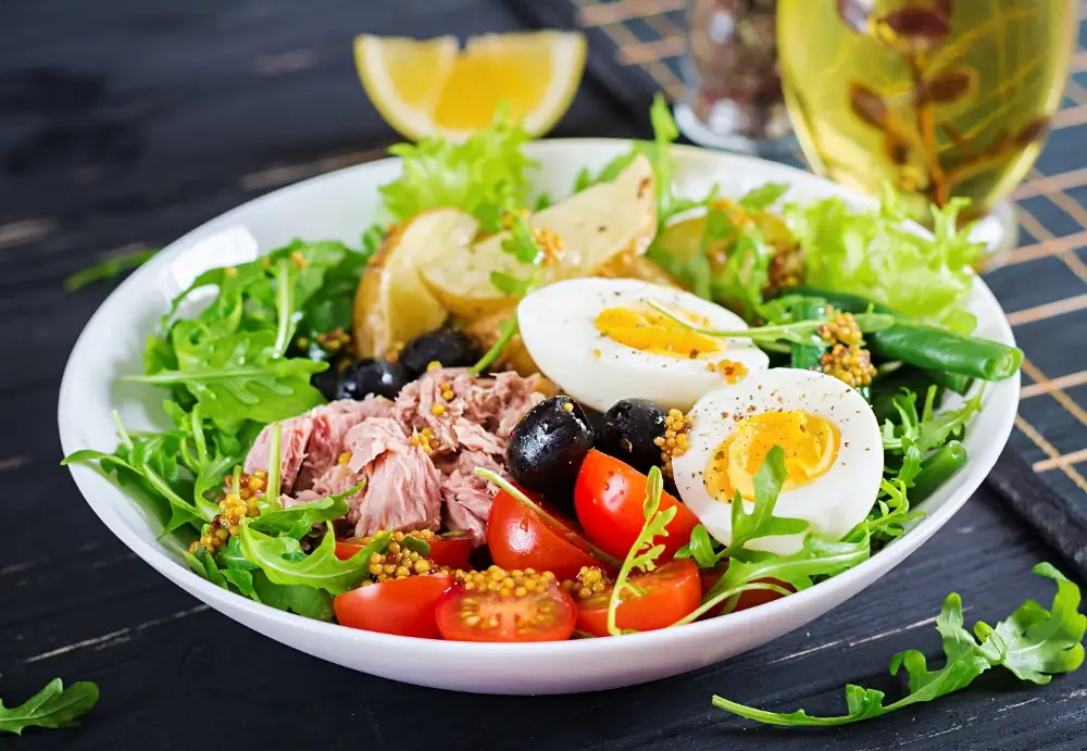 A vibrant protein-packed salad featuring tuna, hard-boiled eggs, cherry tomatoes, black olives, arugula, and green beans, garnished with mustard seeds and served with lemon wedges.