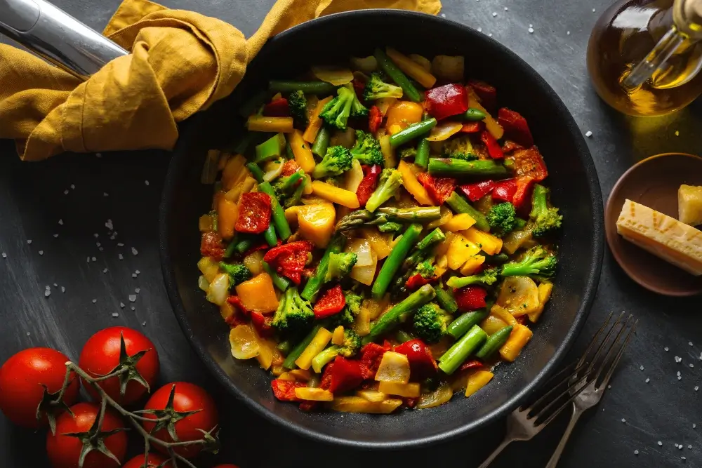 A colorful skillet of stir-fried vegetables including broccoli, green beans, red peppers, and yellow squash, served on a dark table with tomatoes, olive oil, and Parmesan cheese on the side.